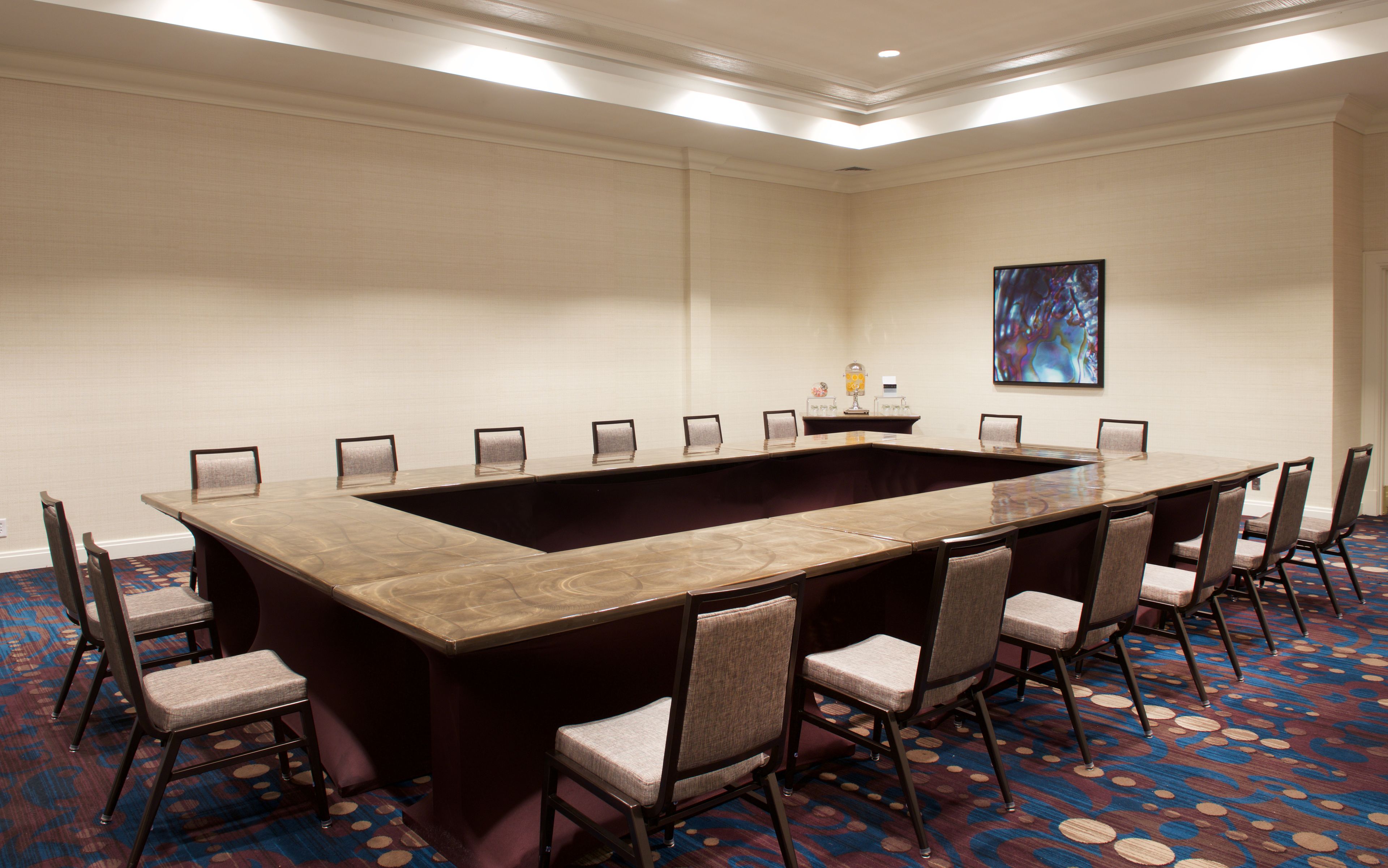 Uma sala de conferência com uma grande mesa retangular rodeada por cadeiras, uma pequena mesa com um abajur e um quadro na parede.