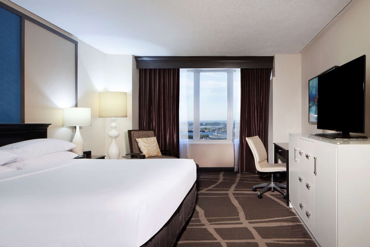 Une chambre d’hôtel avec un grand lit, un bureau avec une chaise, une télévision et une vue sur la ville par la fenêtre.