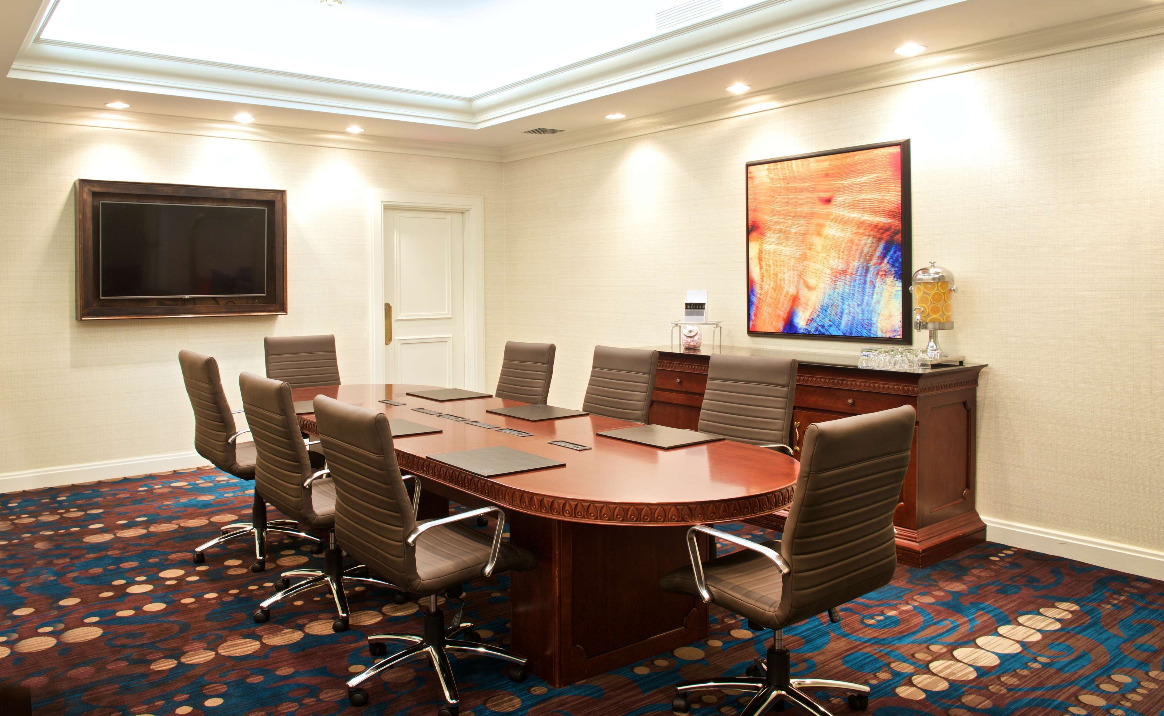 Una sala de conferencias con una gran mesa ovalada, seis sillas, un televisor de pantalla plana, un cuadro decorativo y una nevera pequeña.