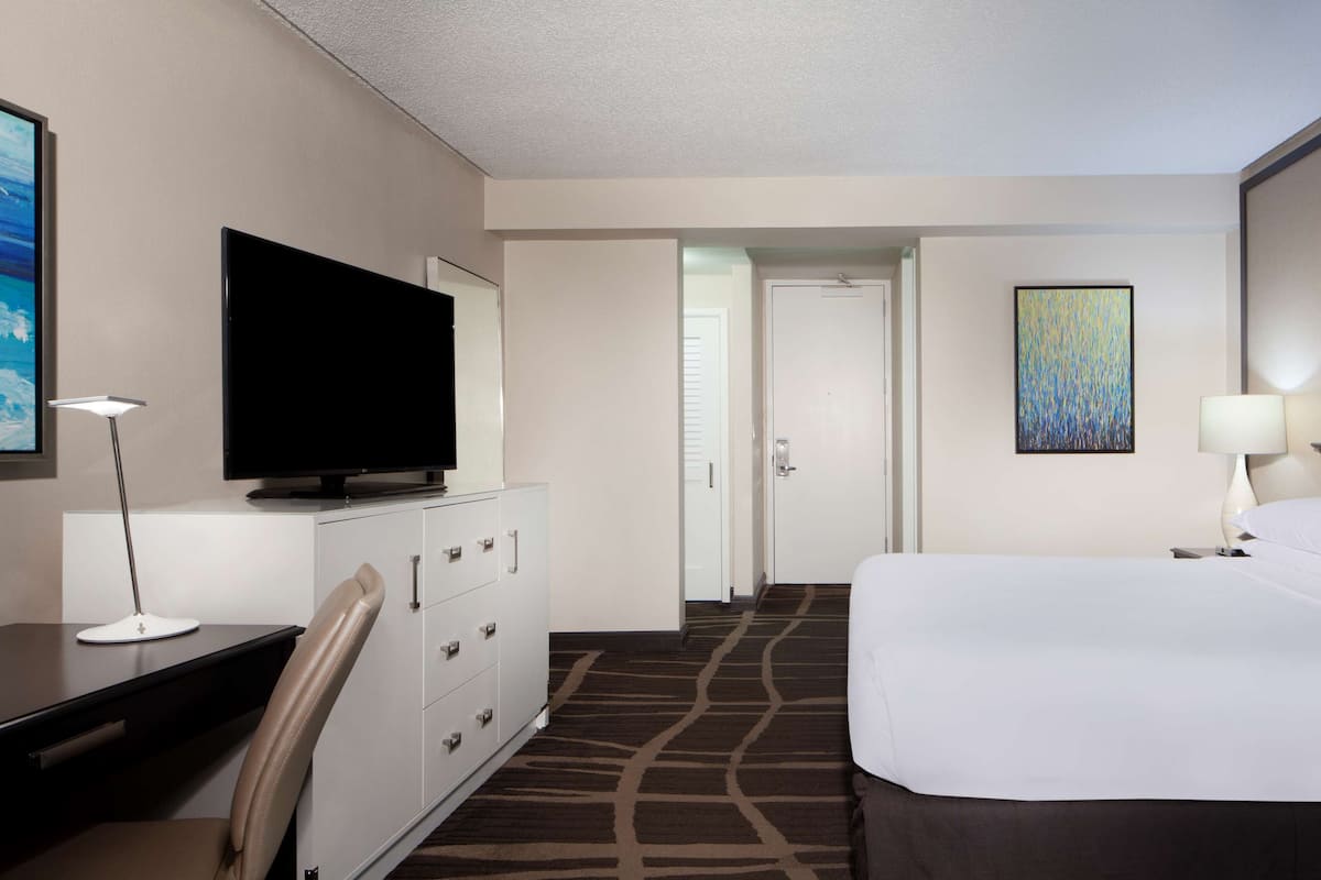 A hotel room with a bed, a desk with a television, a dresser, a lamp, and a painting on the wall.