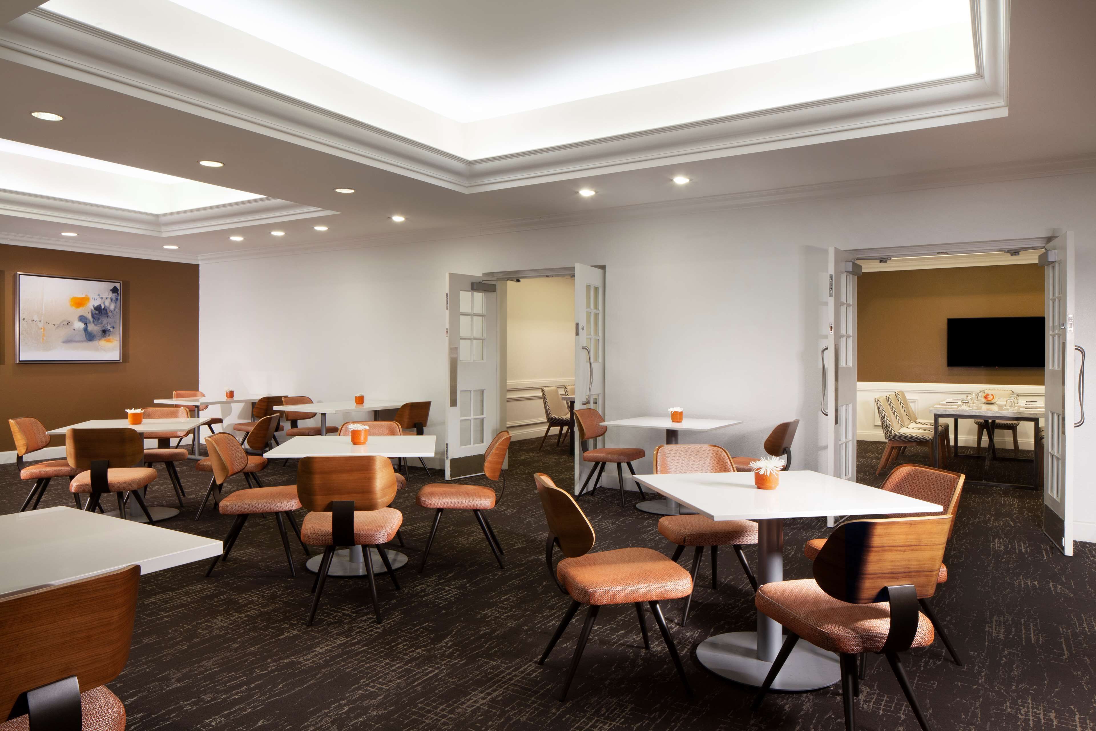 A hotel conference room with multiple tables and chairs, recessed ceiling lights, and a large painting on the wall.