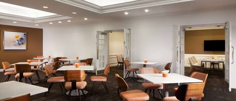 A hotel conference room with multiple tables and chairs, recessed ceiling lights, and a large painting on the wall.