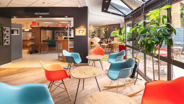 Un café moderno con sillas coloridas, mesas de madera y grandes ventanales.