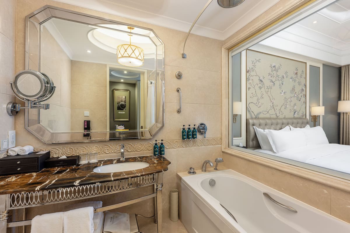 Une salle de bain moderne avec un grand miroir, un lavabo en marbre, une baignoire et un grand lit doté d’une tête de lit.