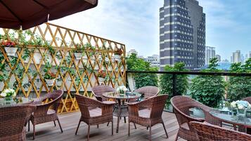 A balcony with wicker furniture and potted plants, overlooking a cityscape with modern buildings.