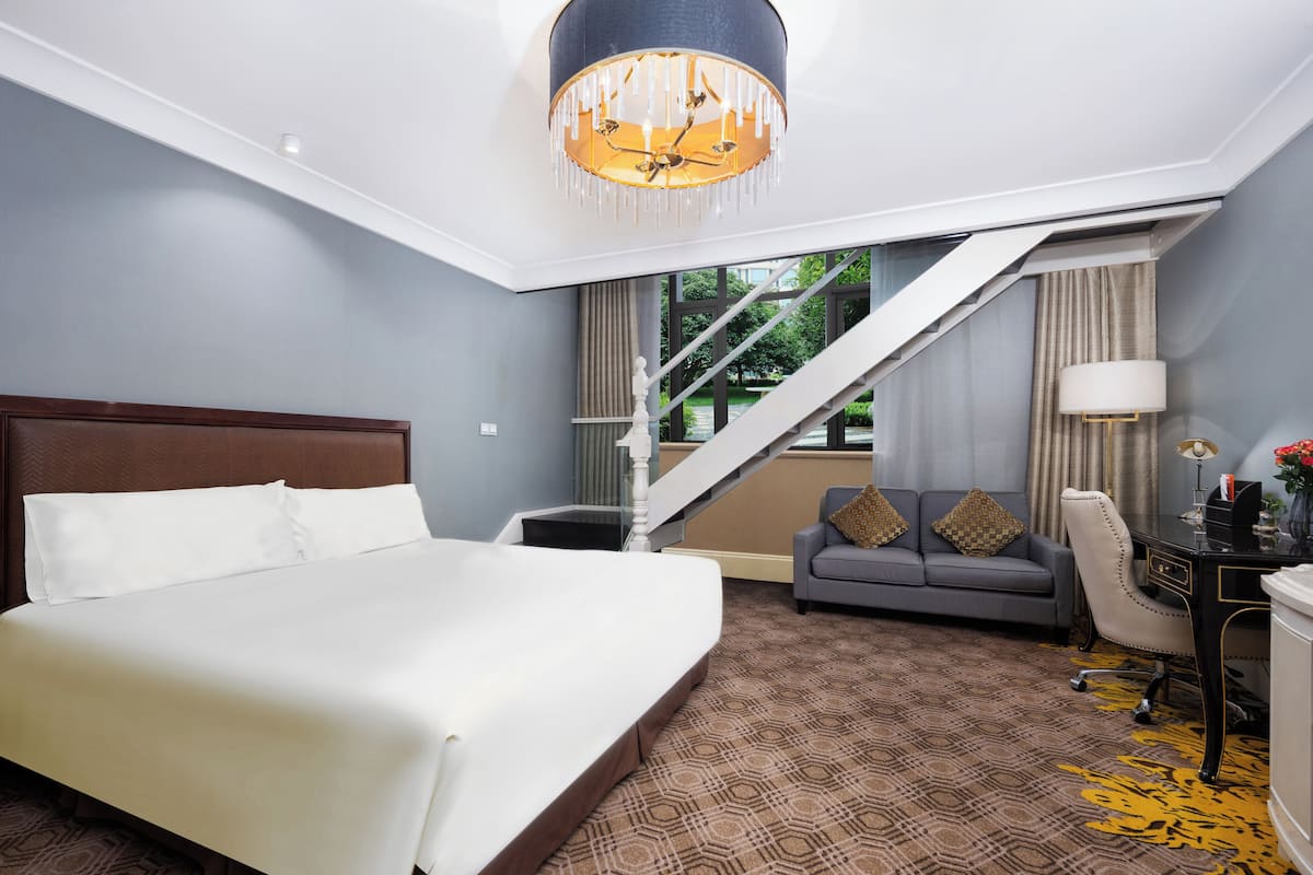 Une chambre d’hôtel avec un grand lit, un canapé gris, un bureau avec une lampe et un escalier menant à un espace extérieur.