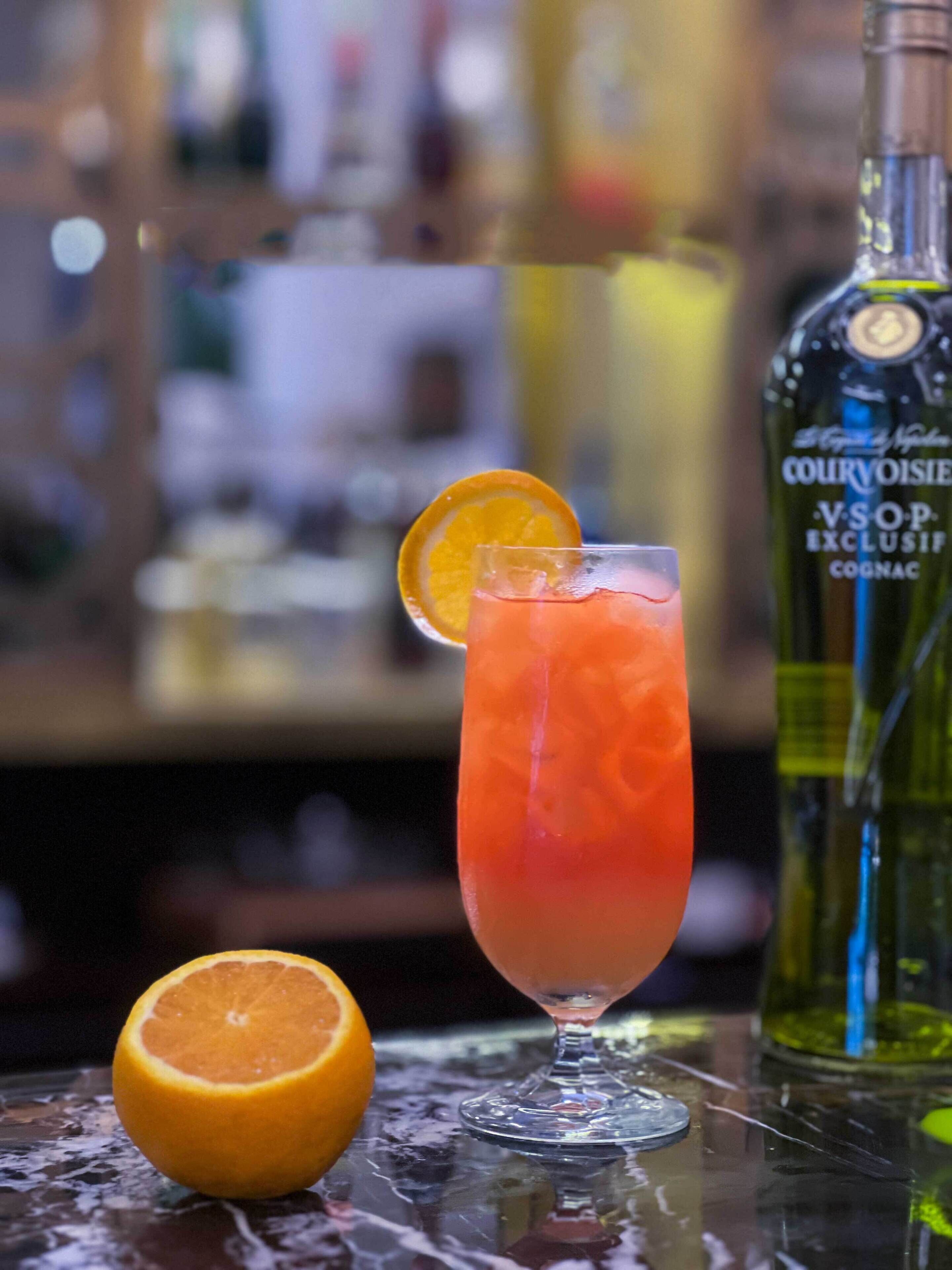 A cocktail glass with a slice of orange on the rim, a bottle of Cognac, and a half orange on a reflective surface.
