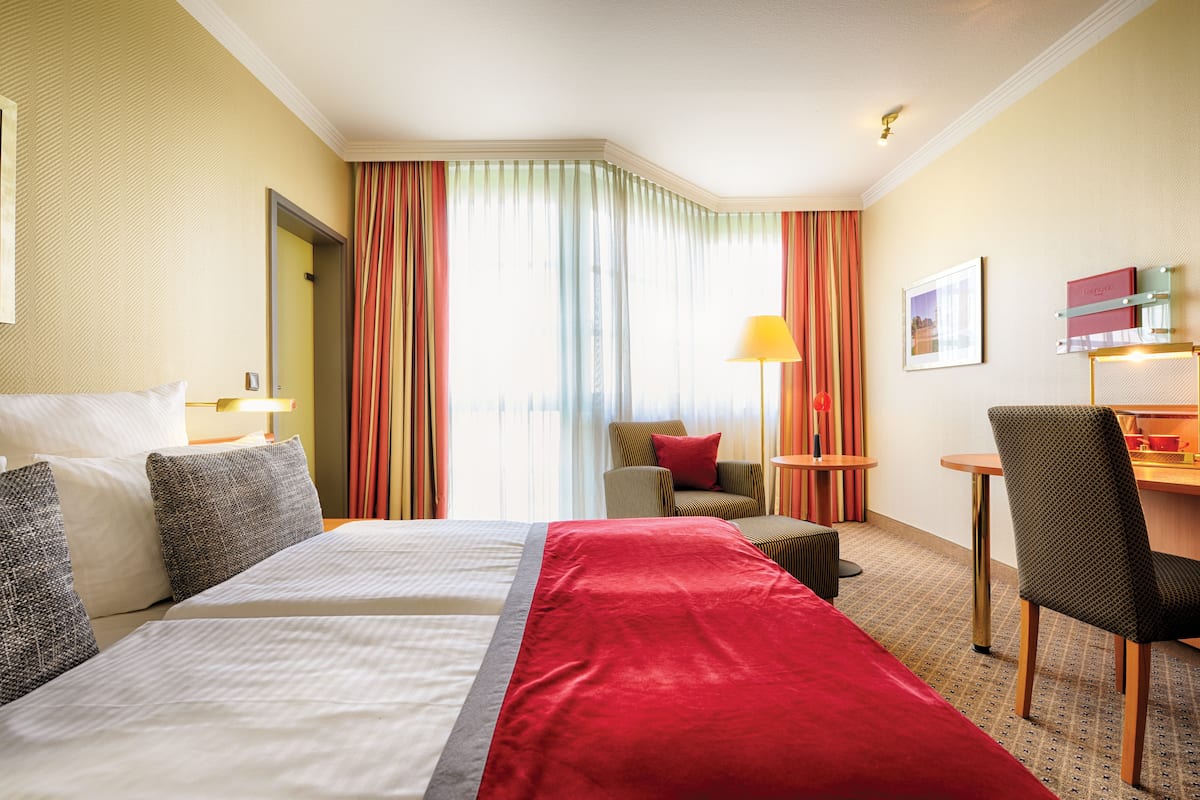 A hotel room with a large bed, a desk, and a chair. There is a window with curtains, a lamp, and a framed picture on the wall.