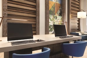 Un espacio de oficina moderno con dos escritorios, cada uno con un ordenador y teclado. Hay dos sillas azules frente a los escritorios. La habitación tiene paneles de madera en las paredes y un cuadro abstracto de gran tamaño.