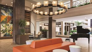 A lobby with a grand piano, orange seating, and a large circular light fixture. - Embassy Suites by Hilton Brea North Orange County (Brea)