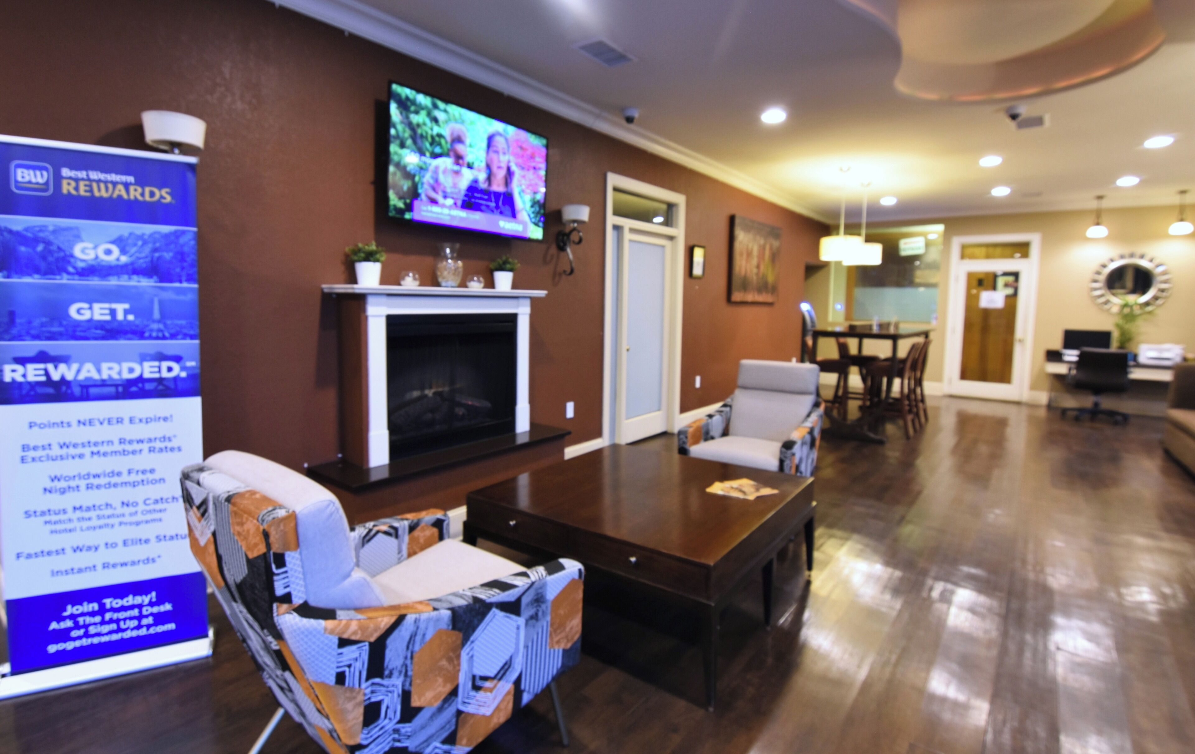 A hotel lobby with a fireplace, a television mounted on the wall, a dining area, and a reception desk.