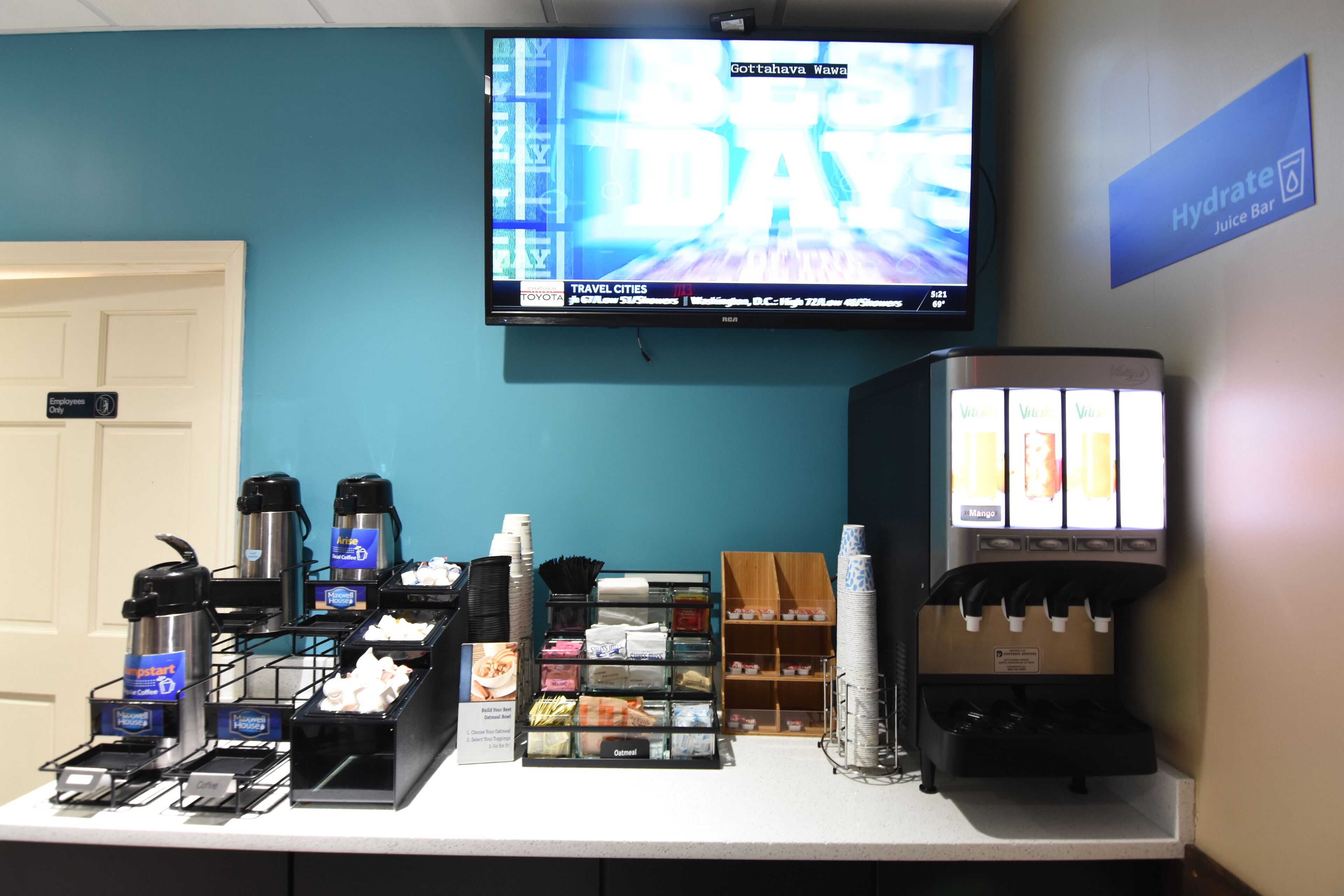 A traveler's lounge with a television displaying a news channel, a hydration station, and a coffee station with various coffee makers and containers.