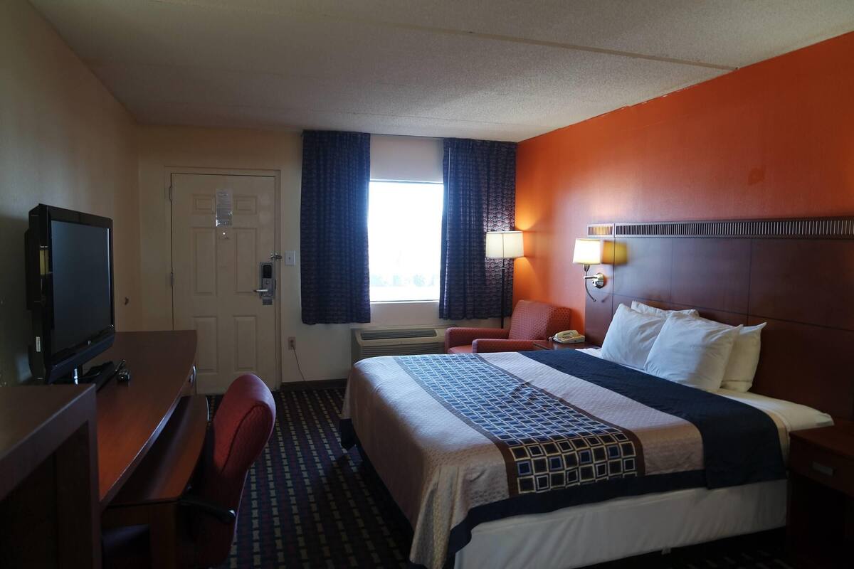 Habitación de hotel con cama, escritorio con televisor, una silla, ventana con cortinas y una lámpara de pared.