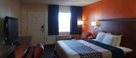 Habitación de hotel con cama, escritorio con televisor, una silla, ventana con cortinas y una lámpara de pared.