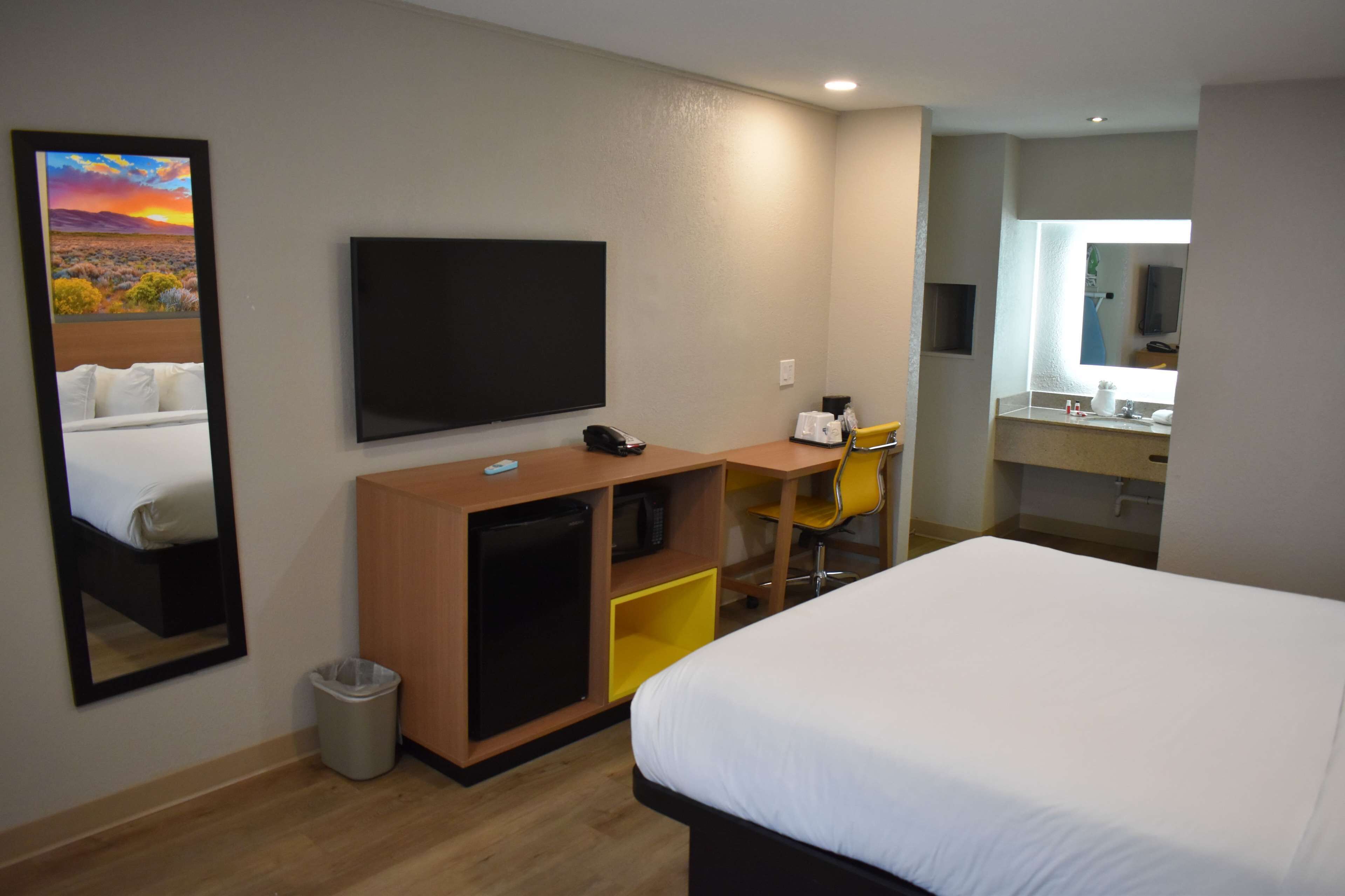 A hotel room with a bed, a desk with a computer, a chair, a TV, a mirror, and a small trash bin.