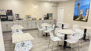 Cocina moderna con cafetera, refrigerador y diversos electrodomésticos. Hay sillas y mesas blancas dispuestas en la habitación.