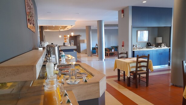 A breakfast buffet area with a variety of food items and beverages, a seating area with tables and chairs, and a kitchen visible in the background. - Flag Hotel Guimaraes Fafe (Fafe)