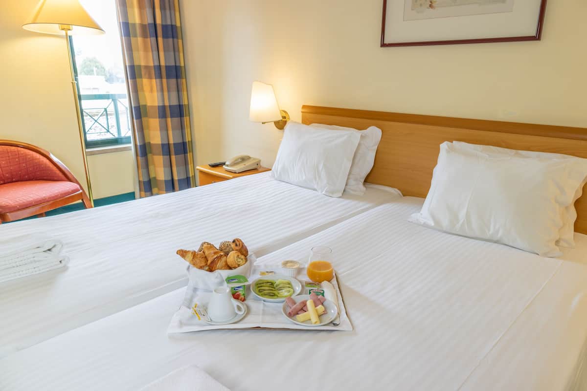 A neatly made bed with white linens, a tray of breakfast including croissants, fruits, and juice, and a red chair in the background.