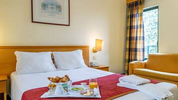 Ein Hotelzimmer mit einem ordentlich bezogenen Bett, einem Frühstückswagen mit Croissants, Früchten, Käse und Saft, einer Nachttischlampe, einem Telefon und einem Fenster mit Vorhängen.