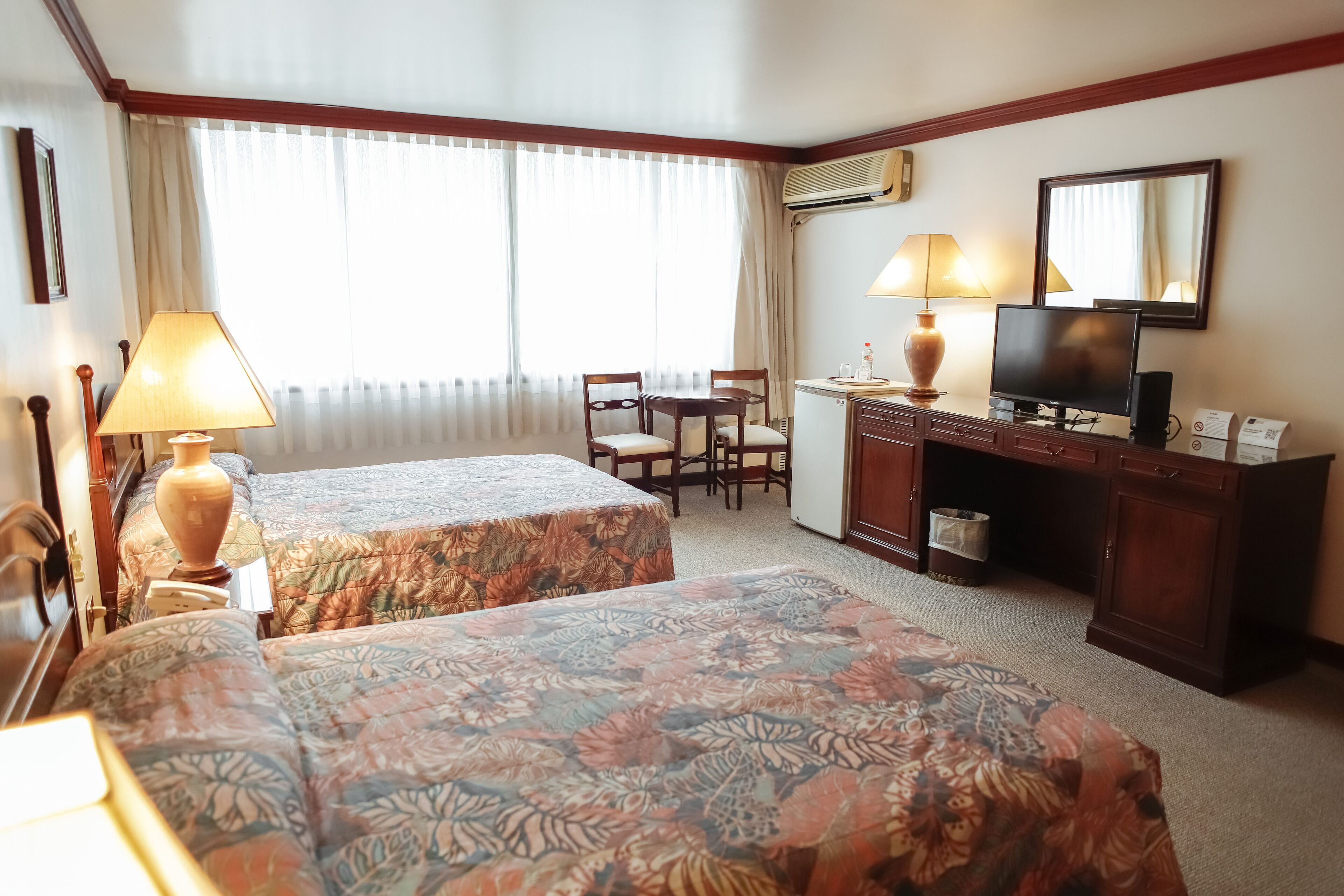 A hotel room with two beds, a desk with a chair, a television, and an air conditioning unit.