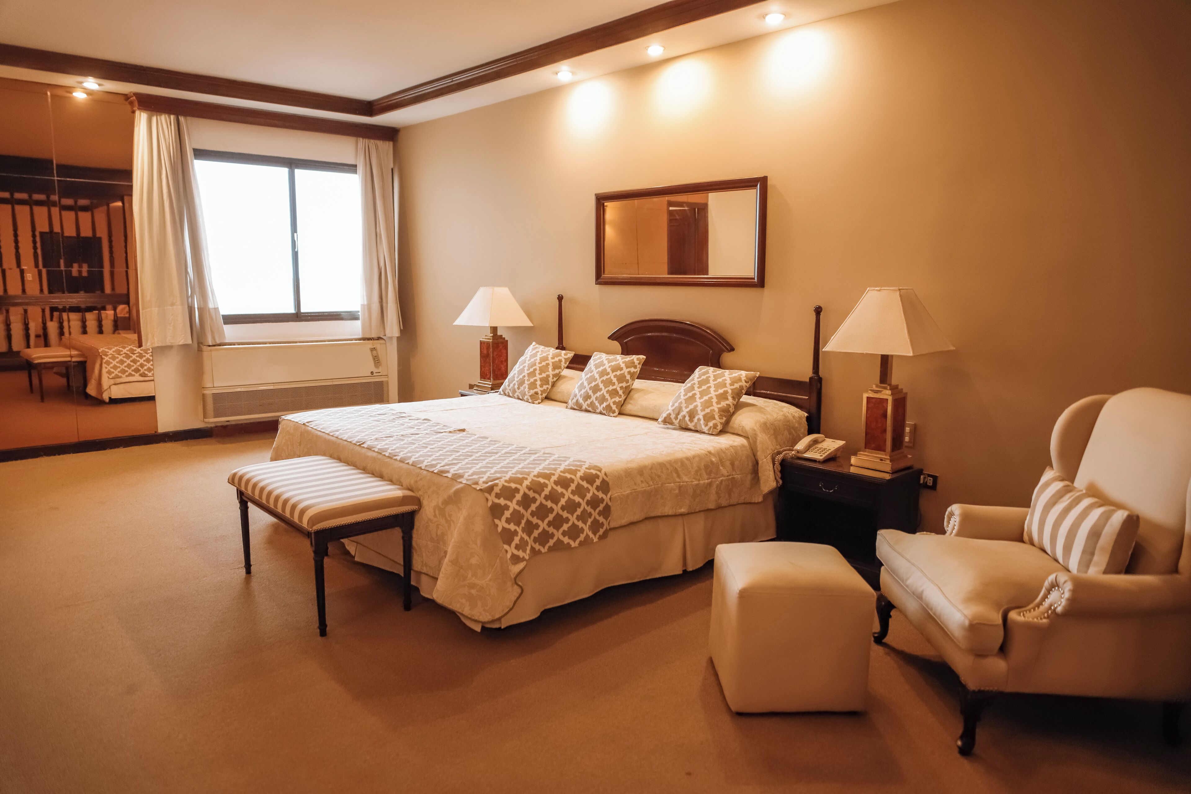 A spacious hotel room with a large bed, a sitting area with a chair and bench, a bedside table with a lamp, and a large mirror on the wall.