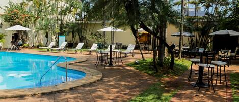 A pool area with lounge chairs and umbrellas, surrounded by palm trees and a building in the background.