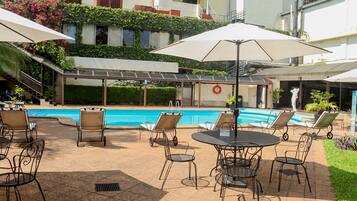 Zona de piscina con sillas de descanso y sombrillas, una piscina y un edificio con una galería techada al fondo.