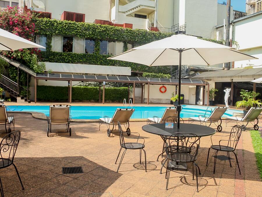Zona de piscina con sillas de descanso y sombrillas, una piscina y un edificio con una galería techada al fondo.