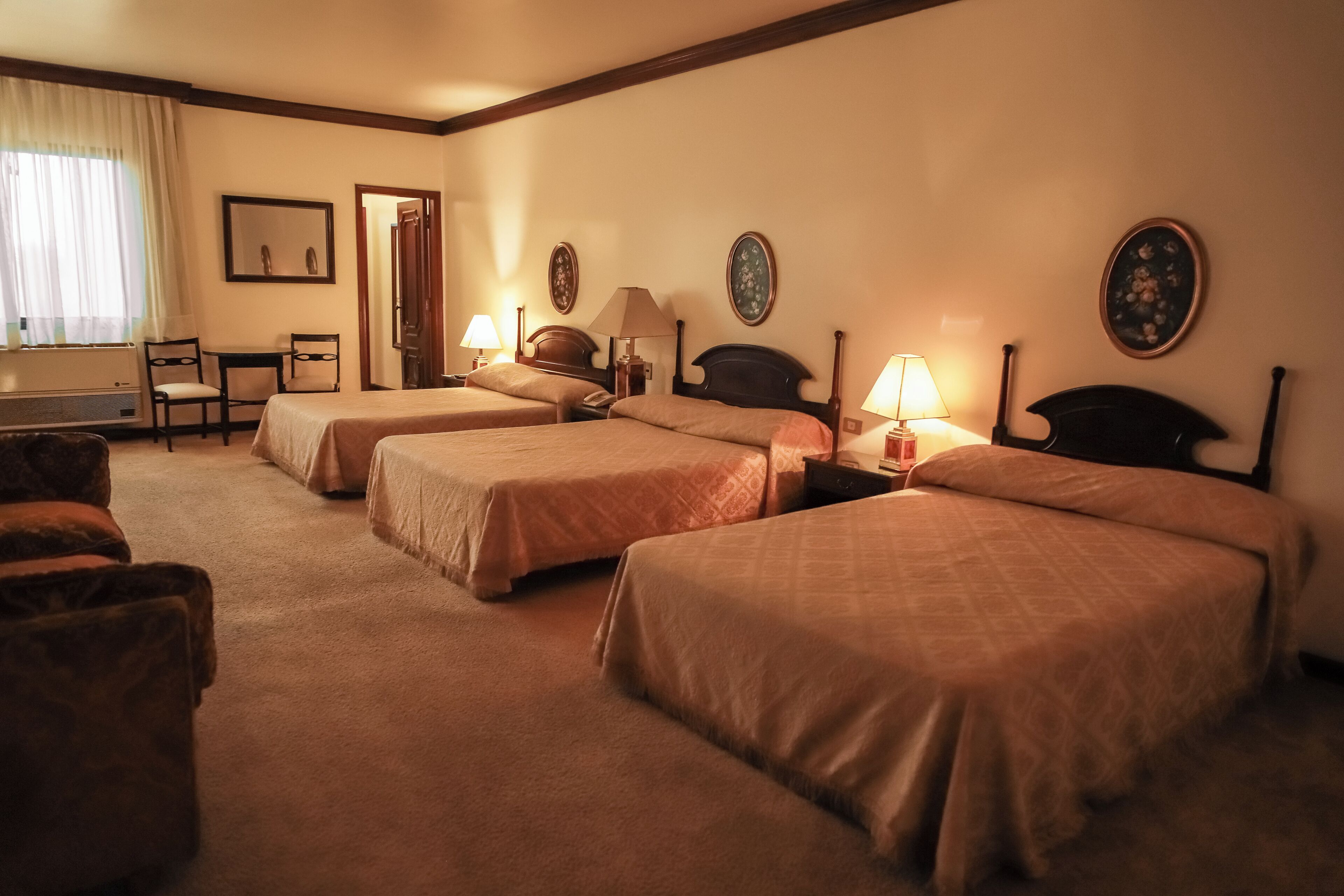 A hotel room with three single beds, a desk with a chair, a small table, and a lamp.