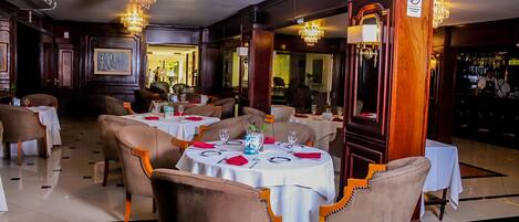 A restaurant with round tables and brown upholstered chairs, chandeliers, and a sign prohibiting smoking.