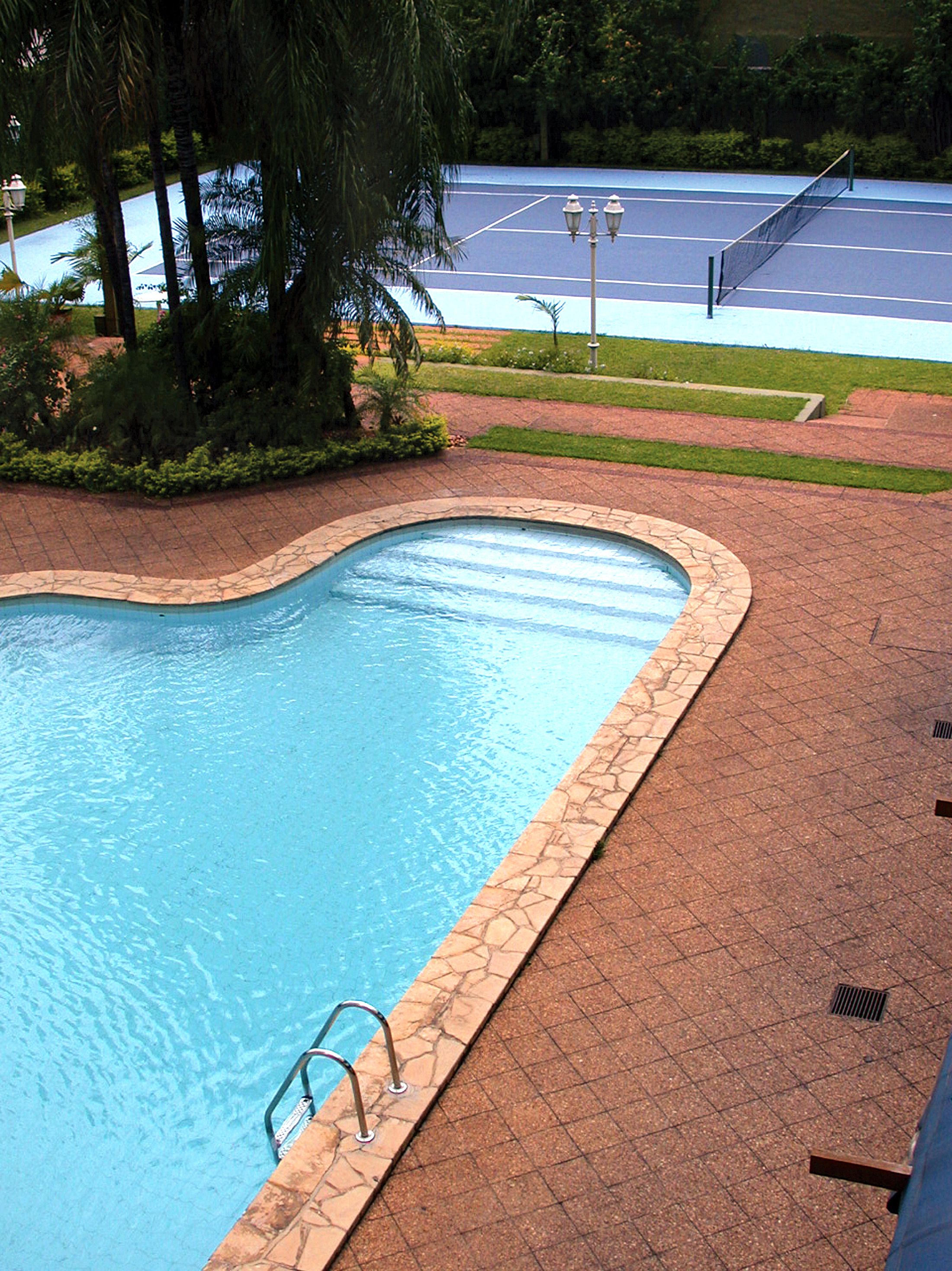 A swimming pool with a stone border, a metal ladder, and a tennis court in the background.