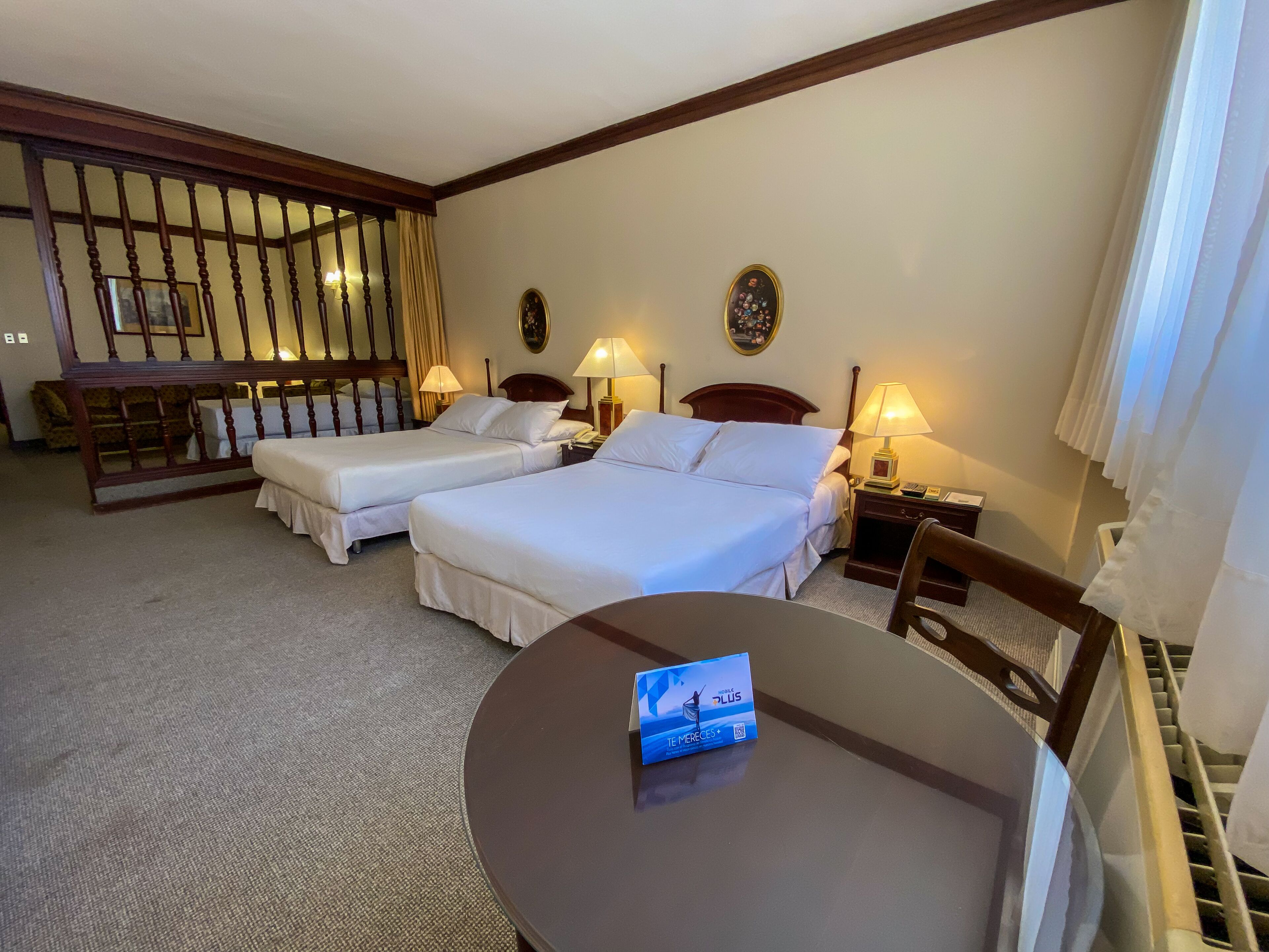 Una habitación de hotel con dos camas individuales, una mesita redonda pequeña y una silla. Hay mesitas de noche con lámparas y una pequeña mesa con un teléfono y un libro.