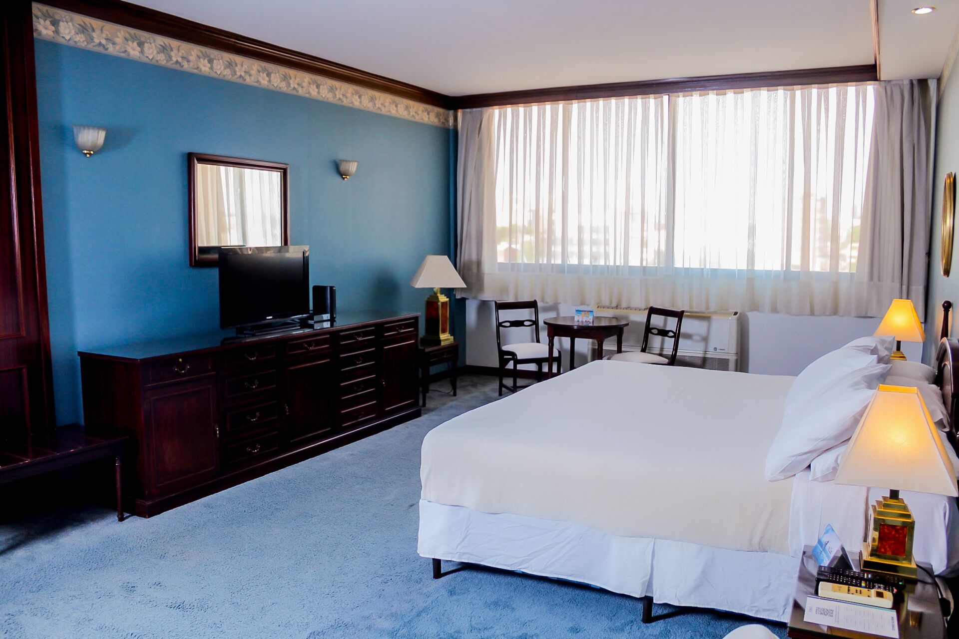 A hotel room with a large bed, a TV on a dresser, a desk with a chair, and a window with curtains.