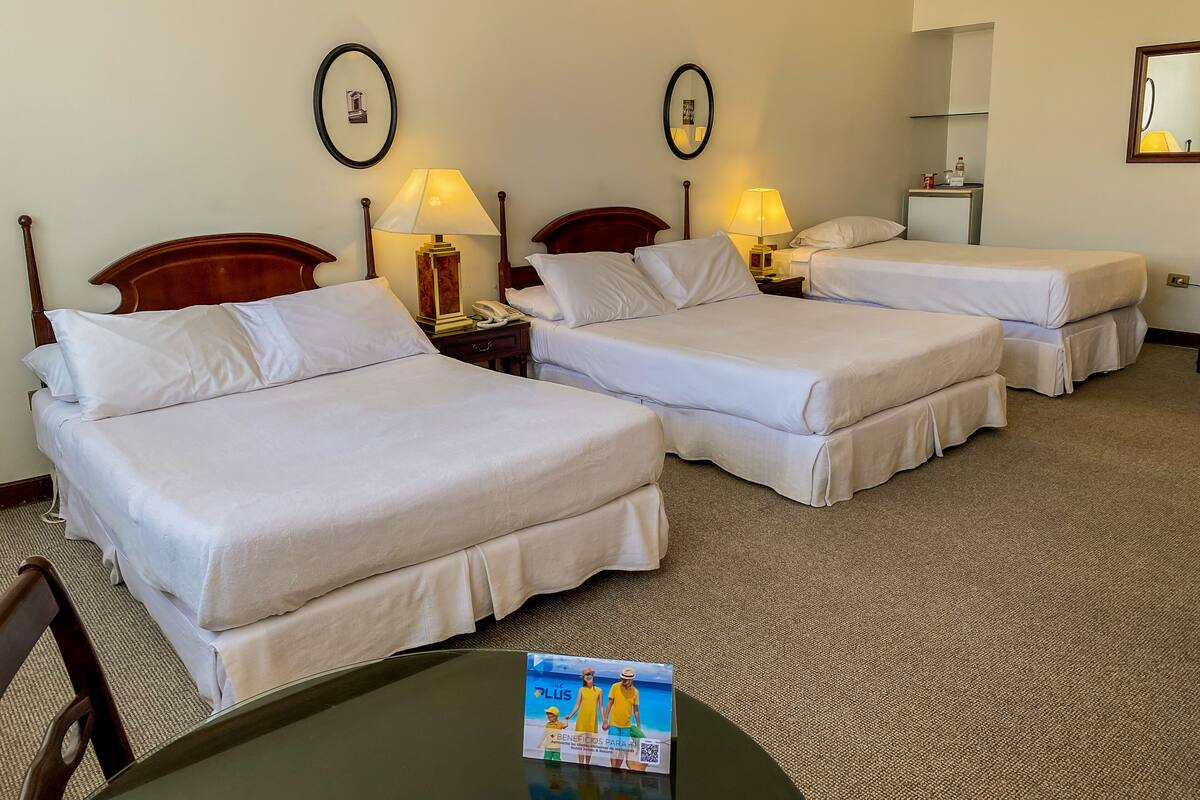 A hotel room with three single beds, a wooden headboard, bedside lamps, a small table with a brochure, and a mirror on the wall.