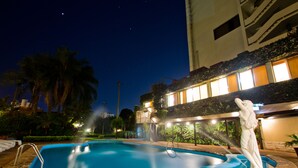 A swimming pool with a statue, surrounded by palm trees and a building with illuminated windows. - Nobile Suites Excelsior Asuncion (Asunción)