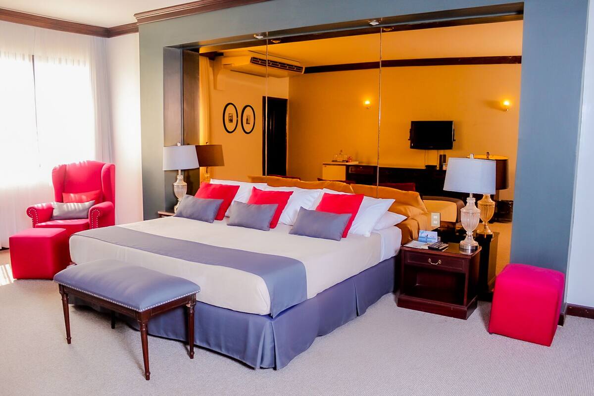 A hotel room with a large bed, a red armchair, a wooden nightstand, and a red ottoman.