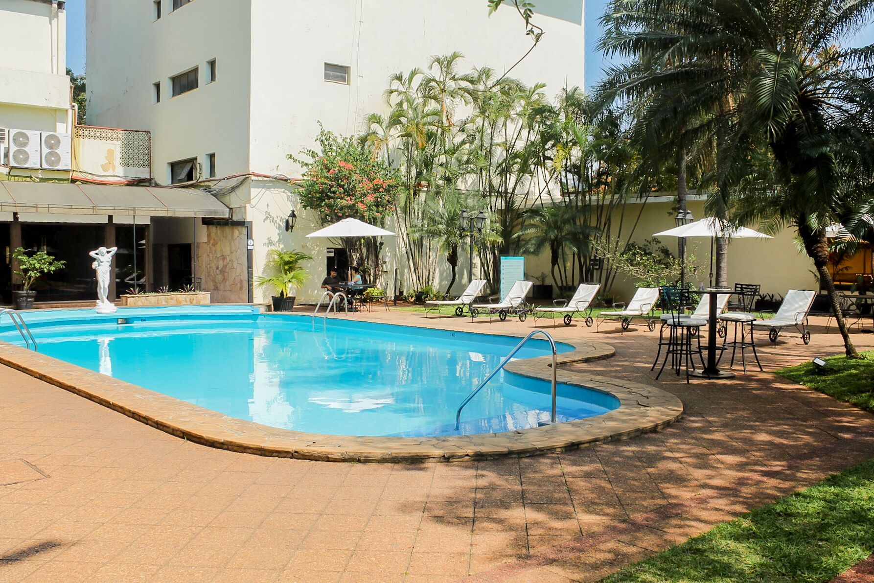A swimming pool with lounge chairs and umbrellas, surrounded by palm trees and a building with multiple windows.