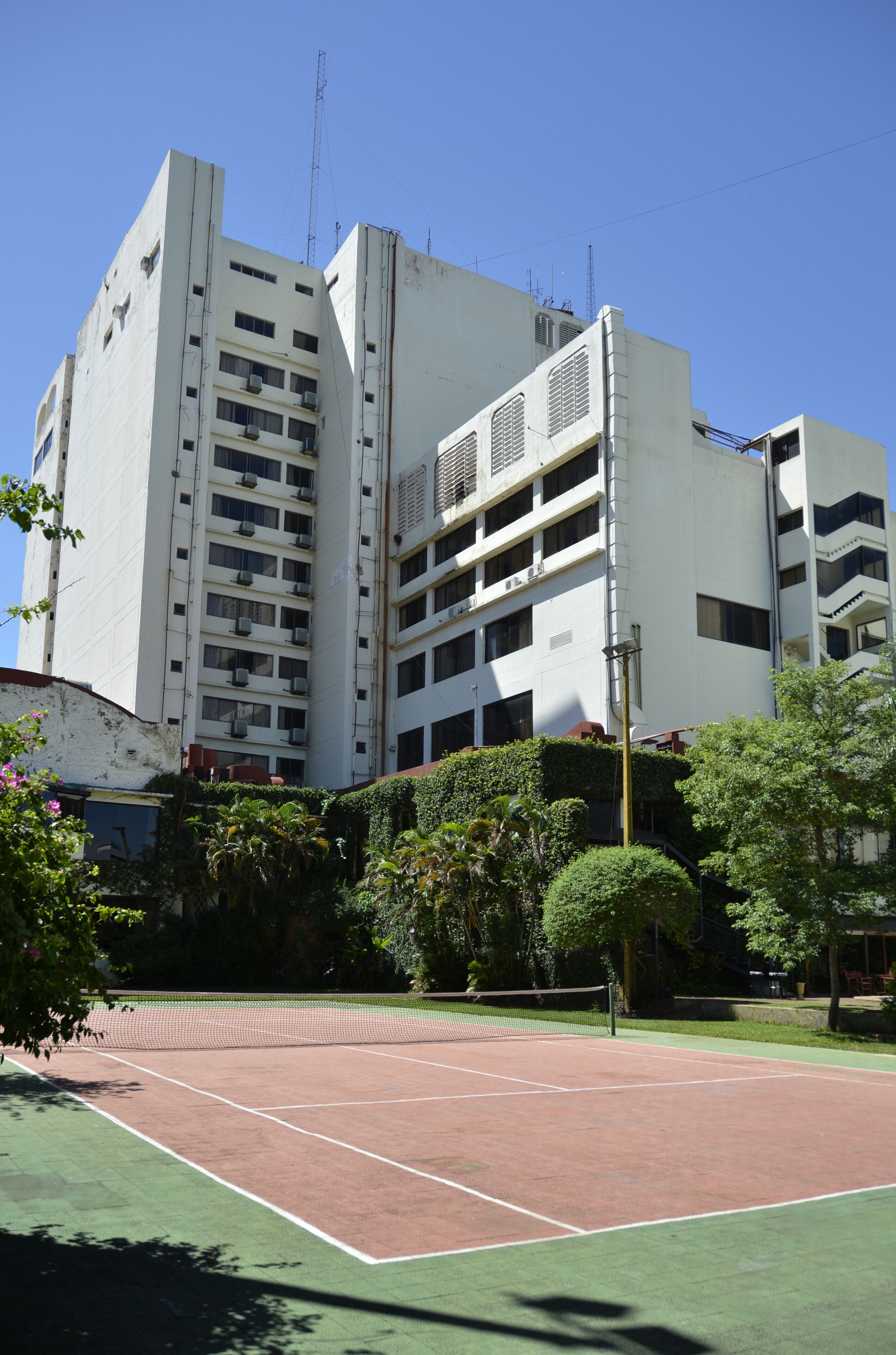 A multi-story building with balconies and air conditioning units, surrounded by greenery and a tennis court.