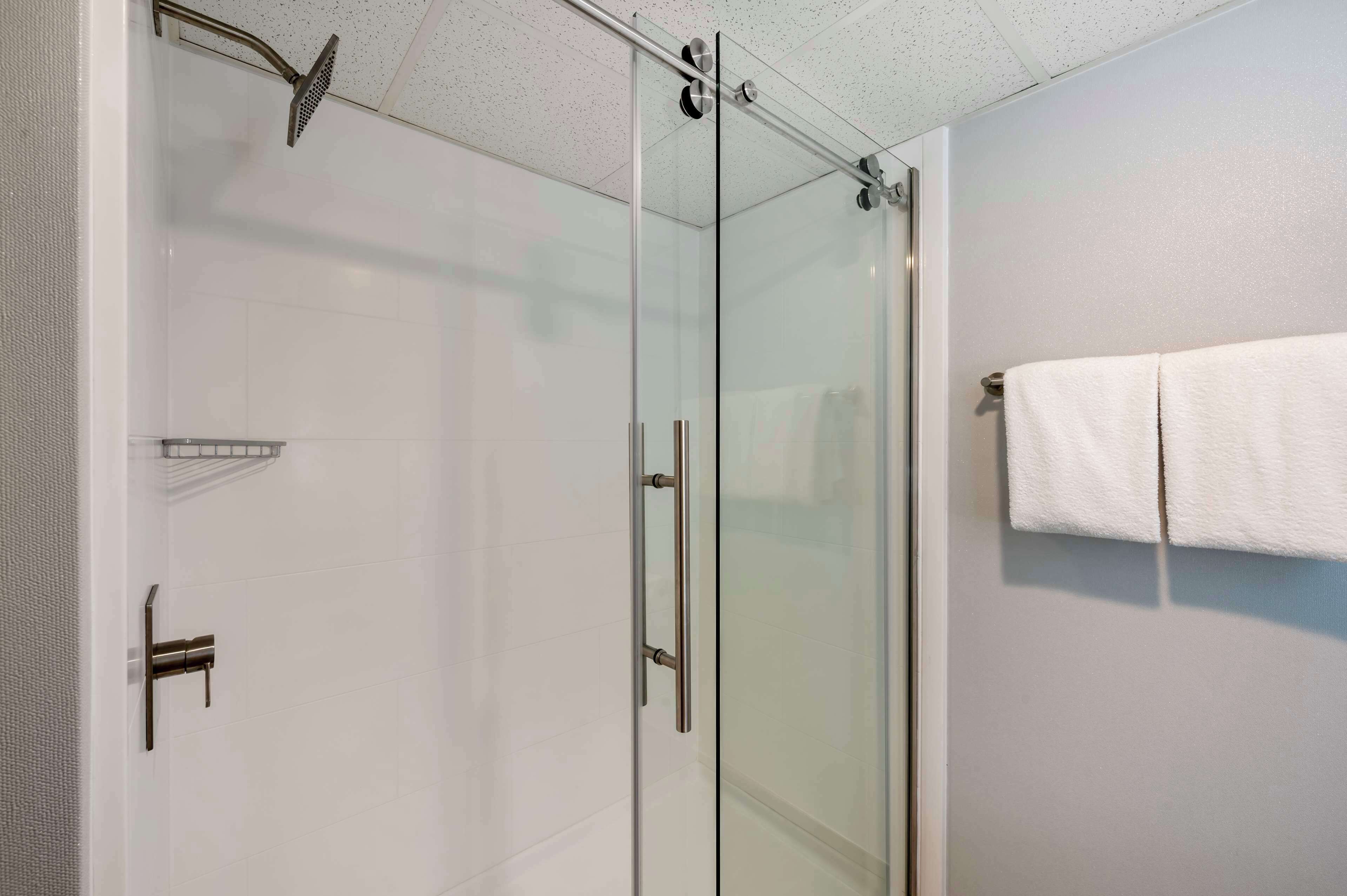 A glass-enclosed shower with a handheld showerhead, a towel rack with a folded towel, and a white tiled wall.