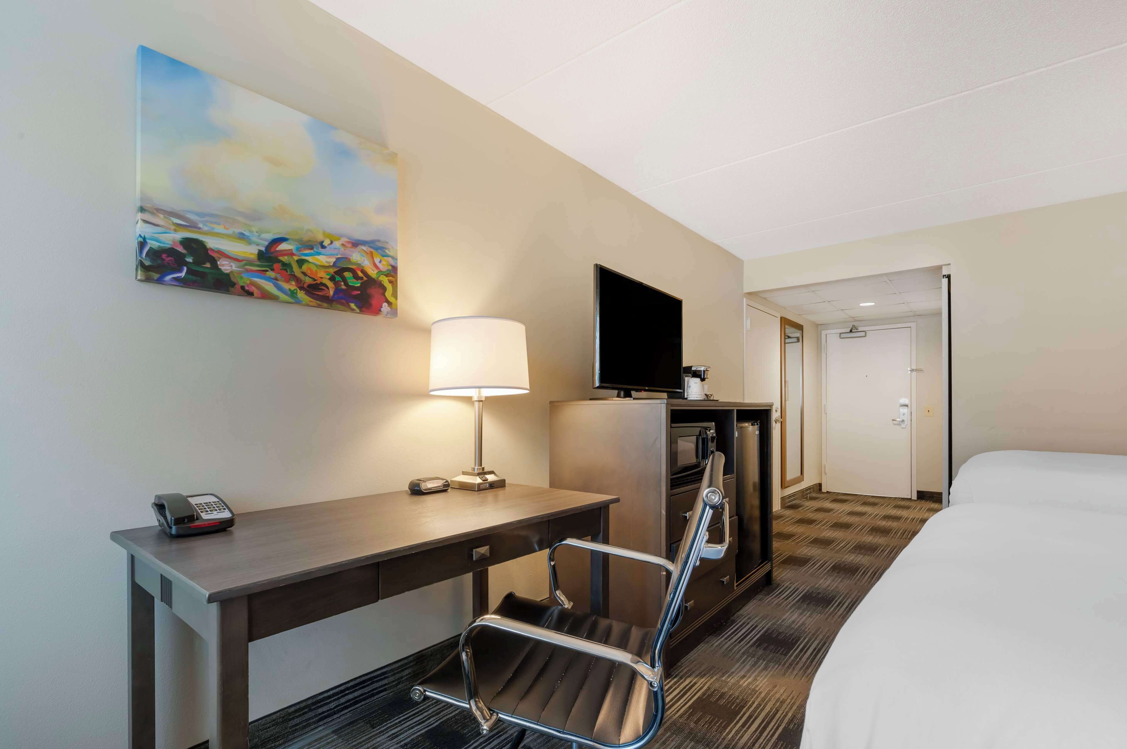 A hotel room with a bed, a desk with a lamp, a chair, a TV, and a painting on the wall.