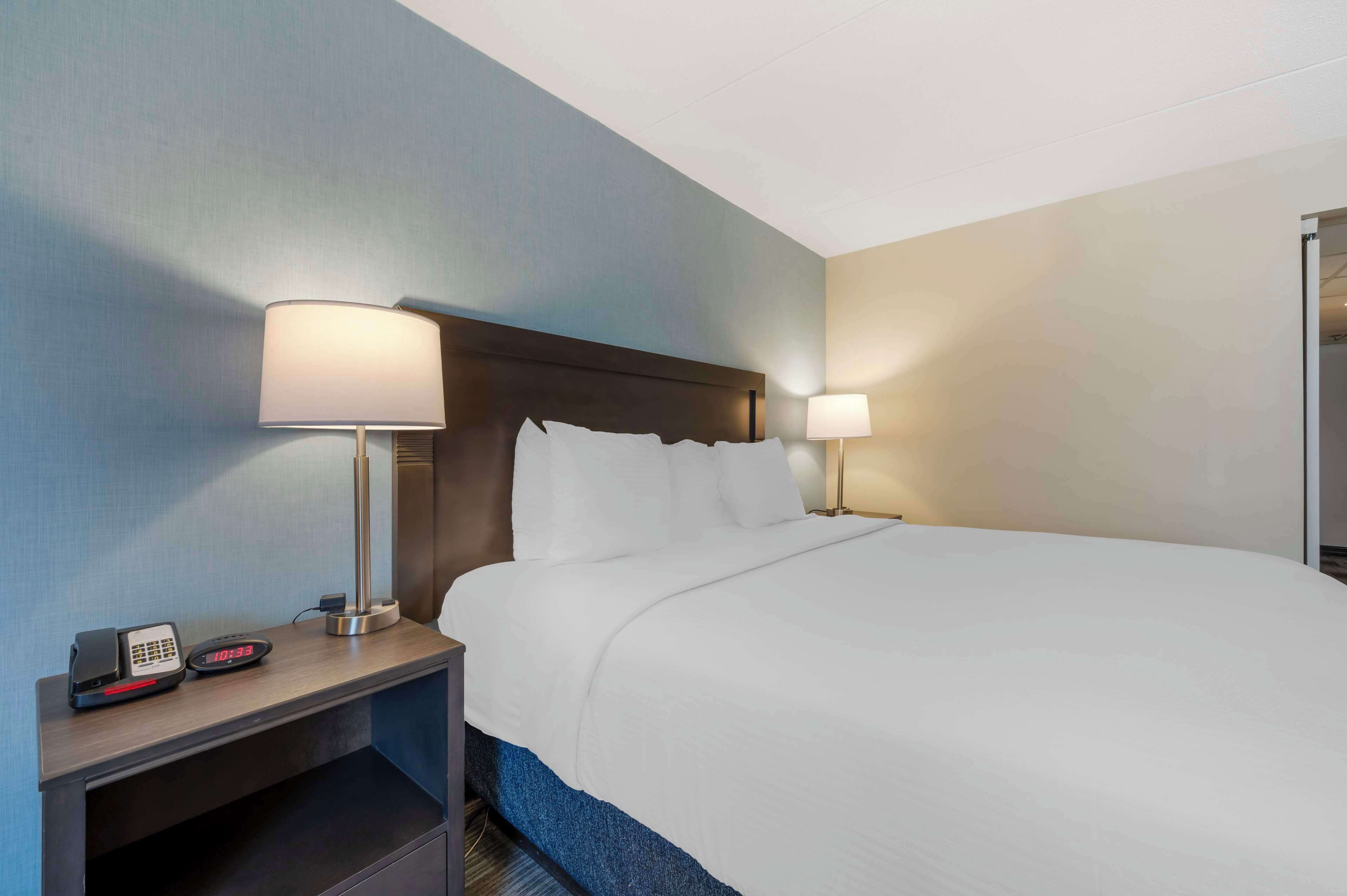 Ein Hotelzimmer mit einem ordentlich bezogenen Bett, zwei Nachttischlampen, einem Nachttisch mit Telefon und digitaler Uhr sowie einem blauen Teppich.