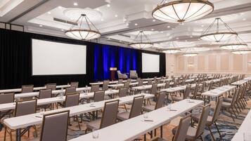 A conference room with rows of tables and chairs, a large screen, and a stage with a blue curtain.