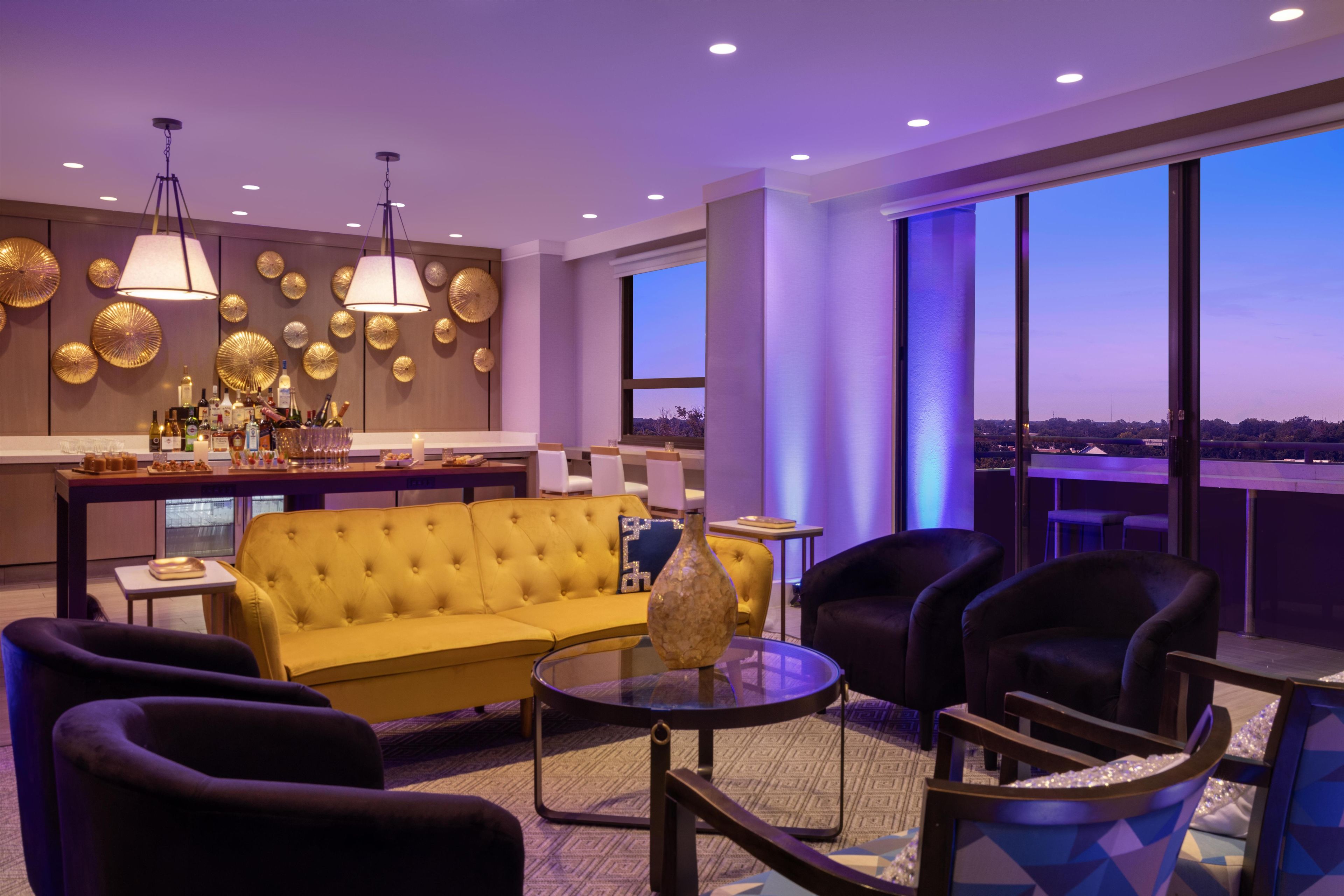 Un moderno vestíbulo de hotel con un sofá amarillo, sillones negros, una mesa redonda y una zona de bar con botellas y vasos.