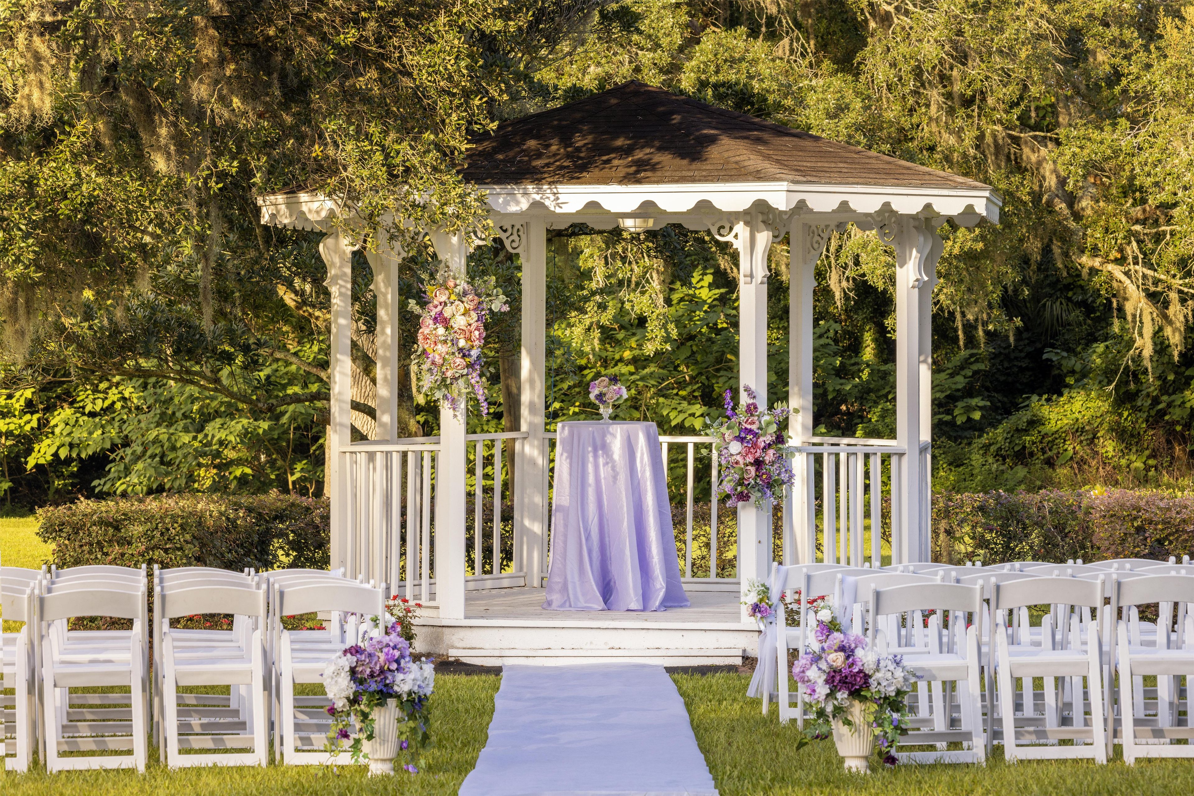 A gazebo with a white exterior and a purple tablecloth, surrounded by white chairs and floral arrangements.