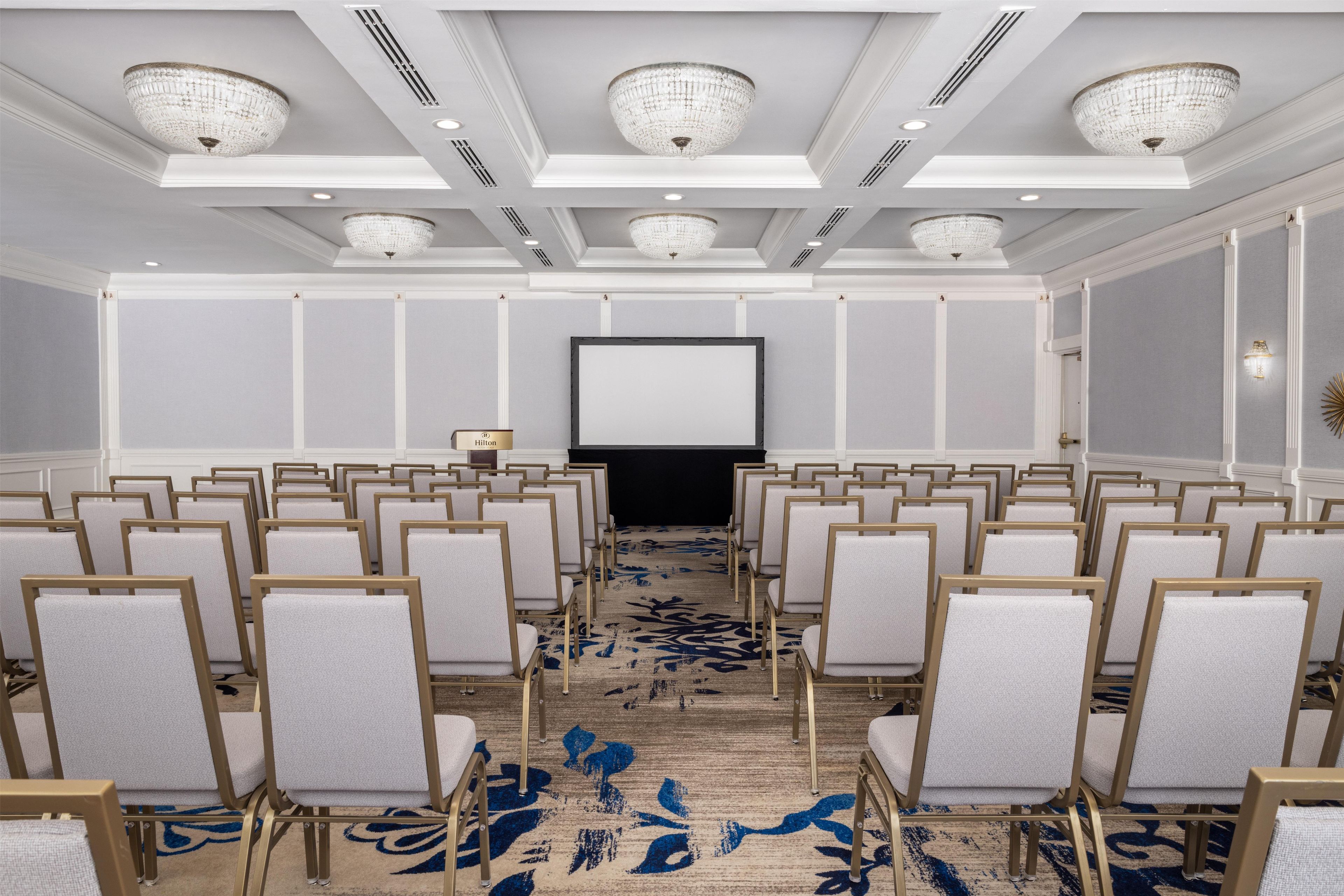 A conference room with rows of chairs, a projector screen, and chandeliers.