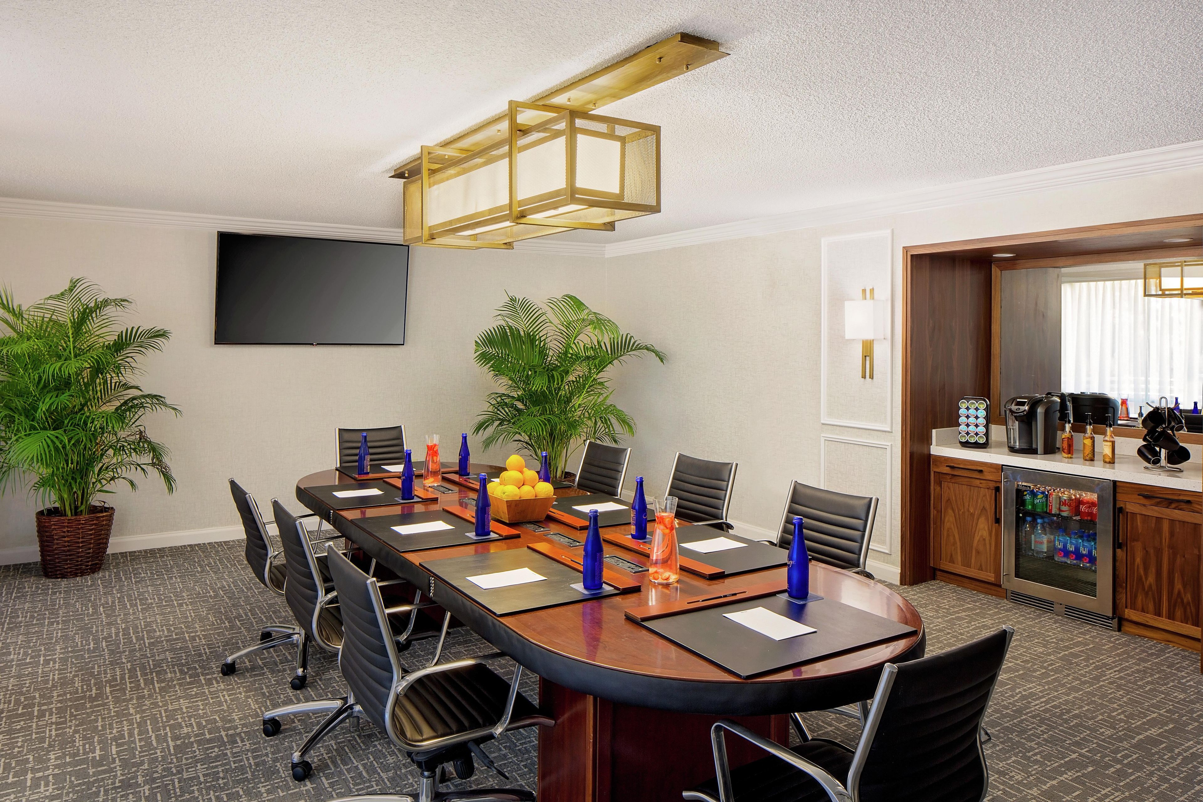 Un salón de conferencias con una mesa larga, sillas, un televisor, plantas y una kitchenette.