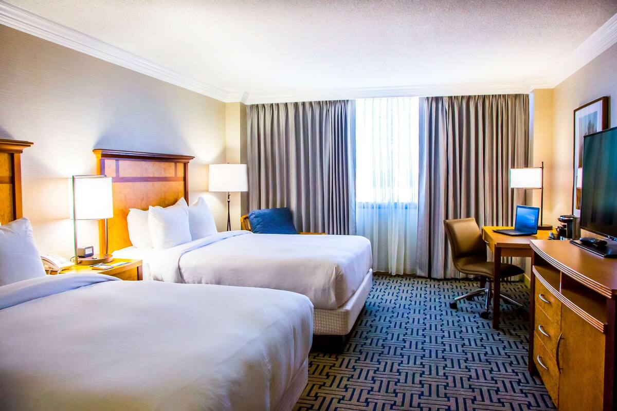 A hotel room with two beds, a desk, a chair, a TV, and a window with curtains.