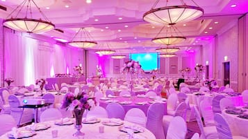 A banquet hall with round tables, white chairs, and chandeliers.