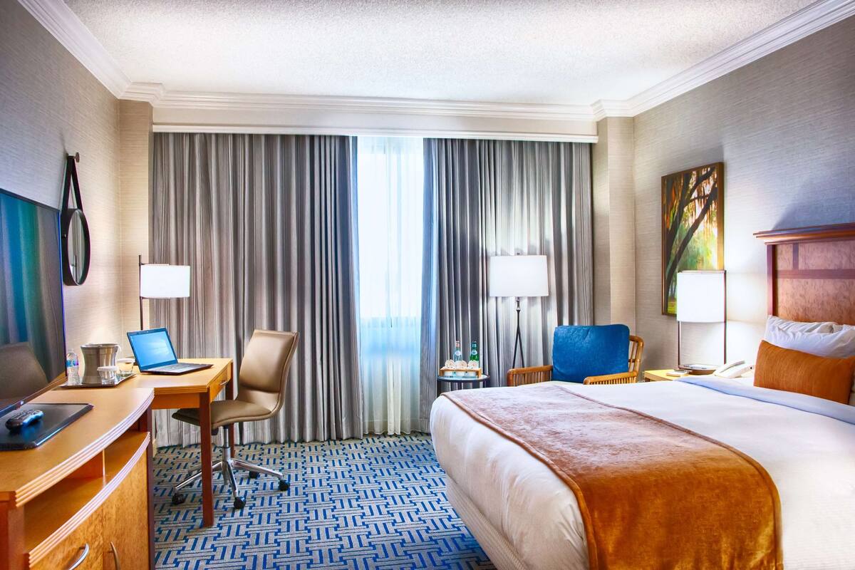 A hotel room with a large bed, a desk with a laptop, a chair, a small table with bottles, and a painting on the wall.