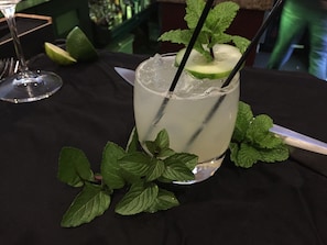 Un verre d’une boisson claire, agrémentée de feuilles de menthe et d’une tranche de citron vert, accompagné d’un couteau et d’une fourchette sur une nappe sombre.